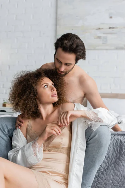 Мужчина без рубашки в джинсах сидит на кровати рядом с кудрявой женщиной в ночном платье и шелковом халате — стоковое фото