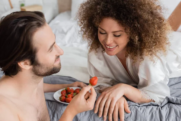 Shirtless man holding ripe strawberry near cheerful young woman in silk robe — Foto stock