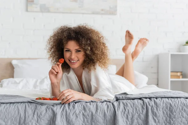 Cheerful young woman in silk robe eating ripe strawberry on bed — Foto stock
