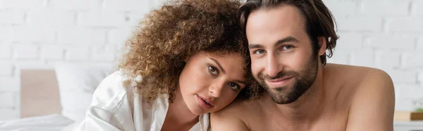 Curly woman in silk robe leaning on shirtless boyfriend smiling in bedroom, banner — Foto stock