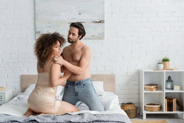 Shirtless man in jeans seducing tattooed young woman in night dress sitting on bed - foto de stock