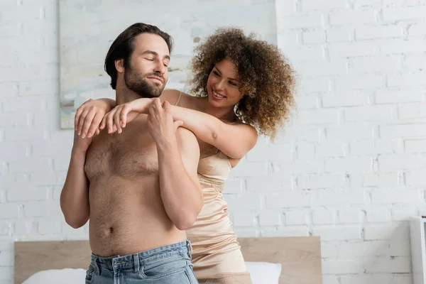 Cheerful and curly young woman hugging shirtless man in denim jeans — Stockfoto
