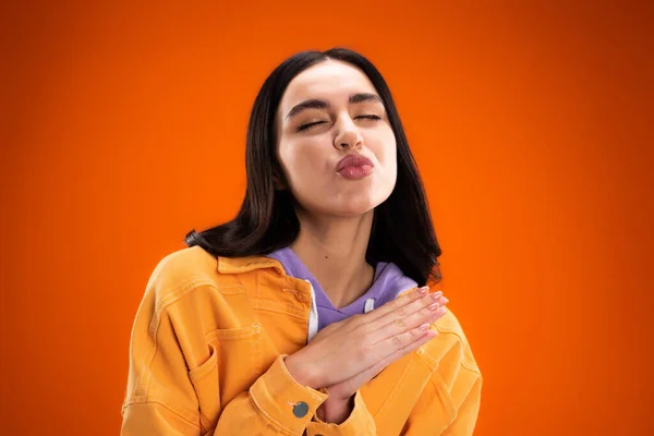 Young brunette woman pouting lips and closing eyes isolated on orange - foto de stock
