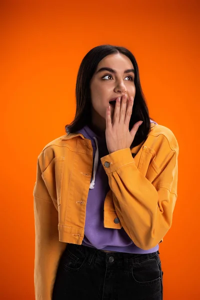 Шокована молода жінка прикриває рот рукою ізольовано на помаранчевому — стокове фото