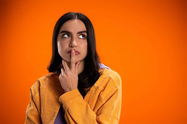 Pensive brunette woman touching lips and looking away isolated on orange — Foto stock
