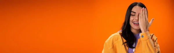 Cheerful brunette woman covering face isolated on orange, banner — Stock Photo