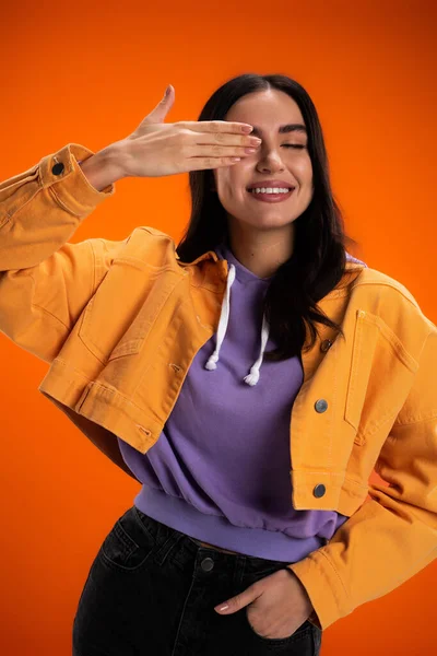 Позитивная женщина в капюшоне и куртке позирует изолированно на оранжевом — стоковое фото