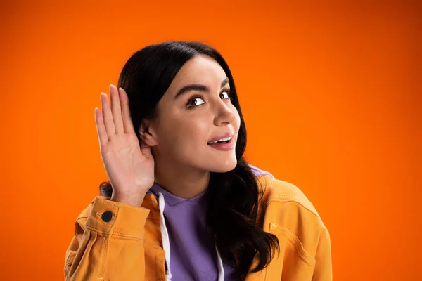 Junge Frau in Jacke hört zu, während sie die Hand am Ohr hält, isoliert auf Orange — Stockfoto