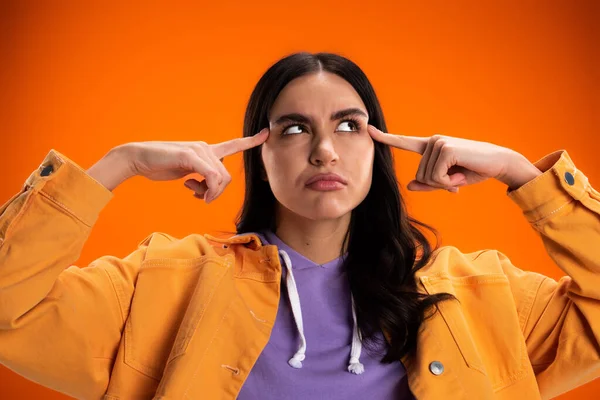 Pensive brunette woman looking away while pointing at head isolated on orange — Stock Photo