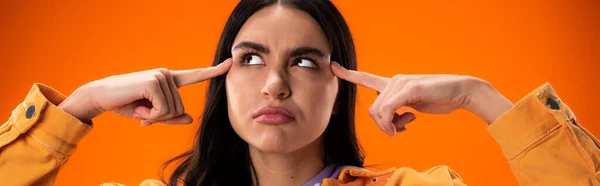 Pensive brunette woman looking away while pointing at head isolated on orange, banner — Foto stock