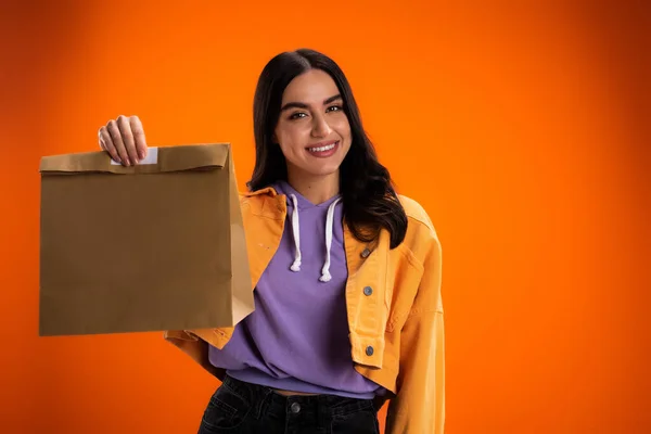Cheerful woman holding paper bag isolated on orange - foto de stock