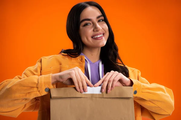 Positive brunette woman holding paper bag and looking at camera isolated on orange — Stockfoto