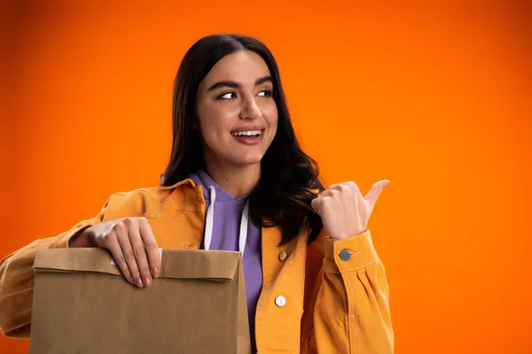 Young woman smiling and pointing with finger while holding paper bag isolated on orange — Photo de stock