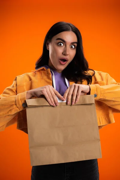 Shocked brunette woman holding paper bag isolated on orange — Stockfoto