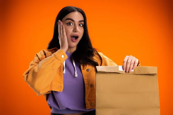 Shocked woman in jacket holding paper bag isolated on orange — Stockfoto