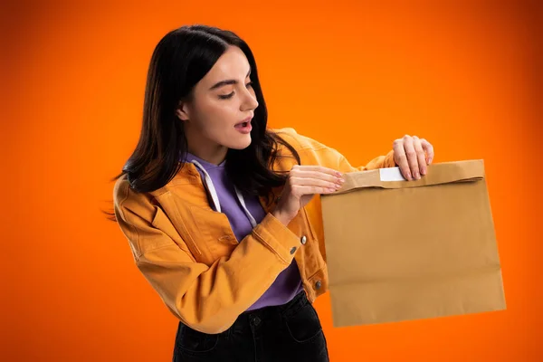Excited brunette woman looking at paper bag isolated on orange — Stockfoto