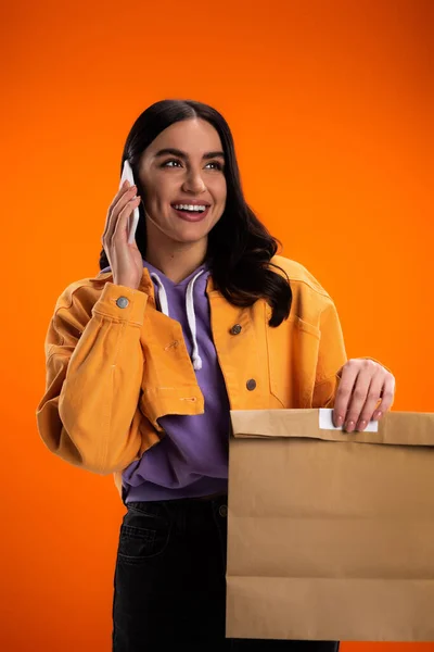 Cheerful brunette woman talking on smartphone and holding paper bag isolated on orange — Photo de stock