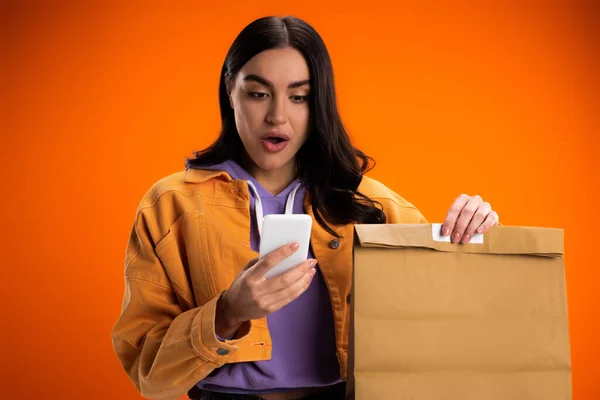 Shocked woman using smartphone and holding takeaway paper bag isolated on orange — Stock Photo