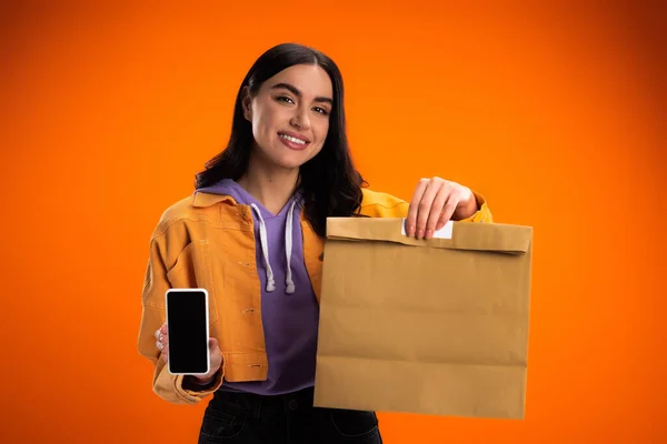 Positive junge Frau zeigt Smartphone und Papiertüte isoliert auf Orange — Stockfoto