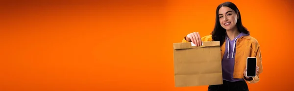 Cheerful woman holding paper bag and smartphone isolated on orange, banner — Stock Photo