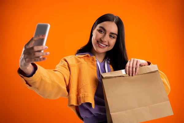 Lächelnde Frau macht Selfie mit Smartphone und hält Papiertüte isoliert auf Orange — Stockfoto