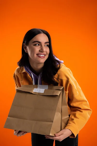 Young brunette woman holding paper bags and looking away isolated on orange — Photo de stock