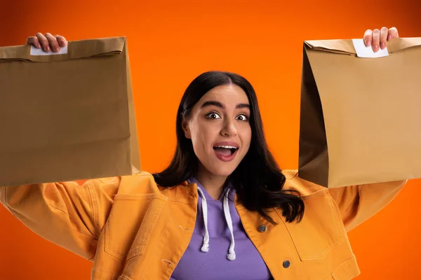 Excited woman in jacket holding paper bags with food isolated on orange — Photo de stock