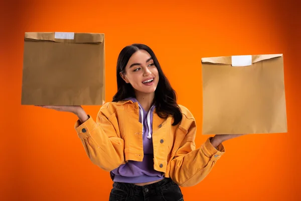Positive young woman holding paper bags from food delivery isolated on orange — Stock Photo