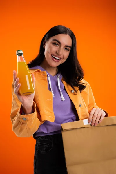 Positive young woman holding bottle with juice and paper bag isolated on orange — Stock Photo