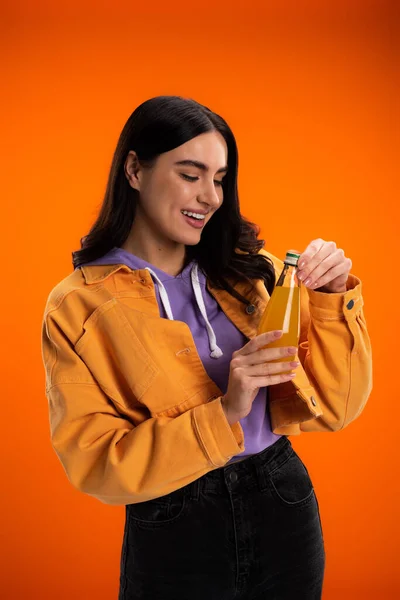 Pretty brunette woman opening bottle with juice isolated on orange — Photo de stock