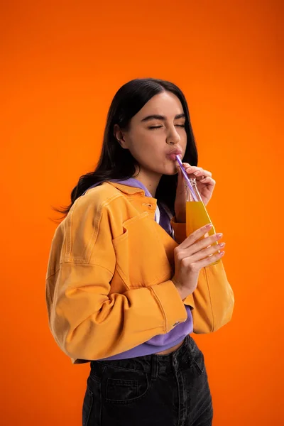 Stylish woman holding glass bottle and drinking juice isolated on orange — Foto stock