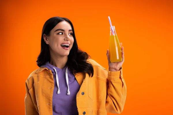 Cheerful brunette woman in hoodie and jacket holding bottle with fresh juice isolated on orange — Photo de stock