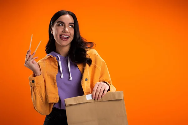 Радісна брюнетка жінка тримає палички і паперовий мішок ізольовано на помаранчевому — стокове фото