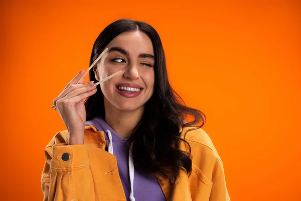 Positive brunette woman holding chopsticks and winking isolated on orange — Photo de stock