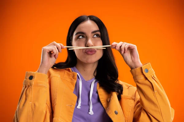 Young woman holding bamboo chopsticks near mouth and pouting lips isolated on orange — Photo de stock