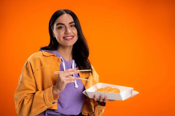Стильная брюнетка смотрит в камеру, держа палочки для еды и суши изолированные на апельсине — стоковое фото