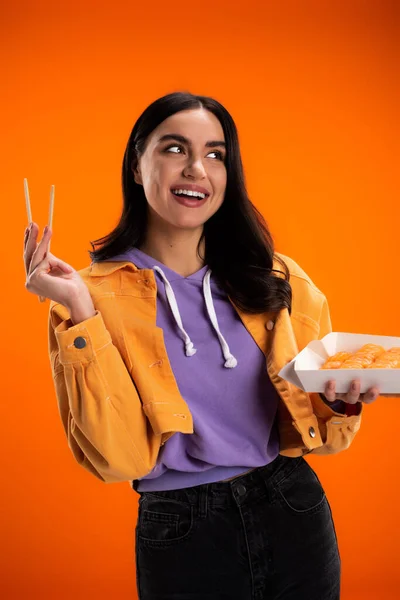 Positive young woman holding chopsticks and takeaway sushi isolated on orange — Stock Photo