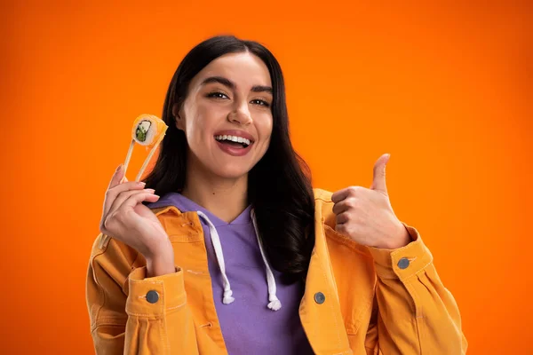 Positive young woman holding sushi in chopsticks and showing like isolated on orange — Stock Photo