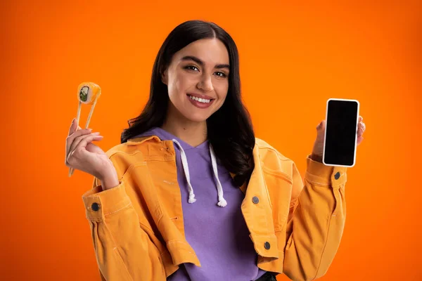 Positive young woman holding sushi in chopsticks and smartphone with blank screen isolated on orange — Fotografia de Stock