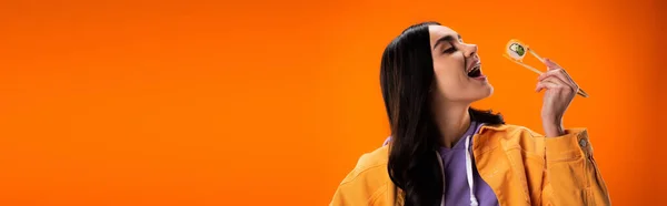 Young woman opening mouth and holding sushi in chopsticks isolated on orange, banner — Foto stock