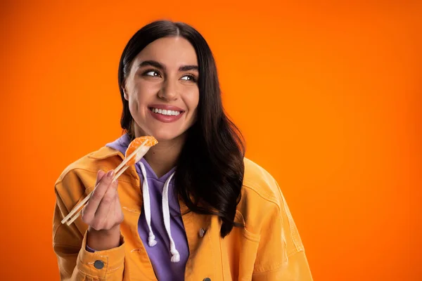 Smiling woman in trendy jacket holding chopsticks with fresh sashimi isolated on orange - foto de stock