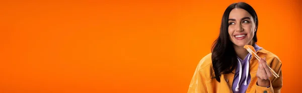 Zufriedene brünette Frau hält Sashimi mit Essstäbchen in der Hand und schaut isoliert auf orangefarbenem Banner weg — Stockfoto