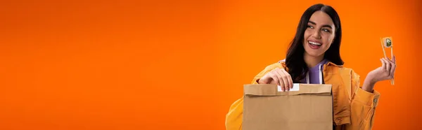 Положительная брюнетка женщина с бумажным пакетом и свежим суши рулон глядя в сторону изолированы на оранжевый, баннер — стоковое фото