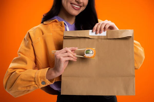 Cropped view of smiling woman with sushi roll and paper bag isolated on orange — Photo de stock