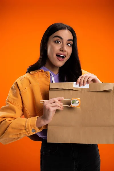 Mujer sorprendida sosteniendo palillos con rollo de sushi cerca de bolsa de papel aislado en naranja - foto de stock