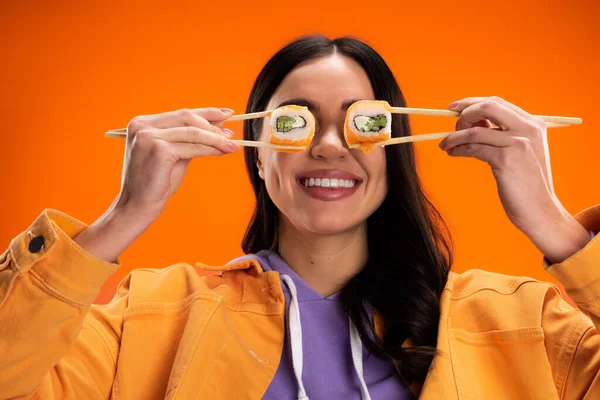 Cheerful woman in jacket and purple hoodie covering eyes with sushi rolls isolated on orange — Stockfoto