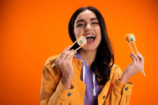 Pleased woman in trendy jacket eating tasty sushi roll and looking at camera isolated on orange - foto de stock