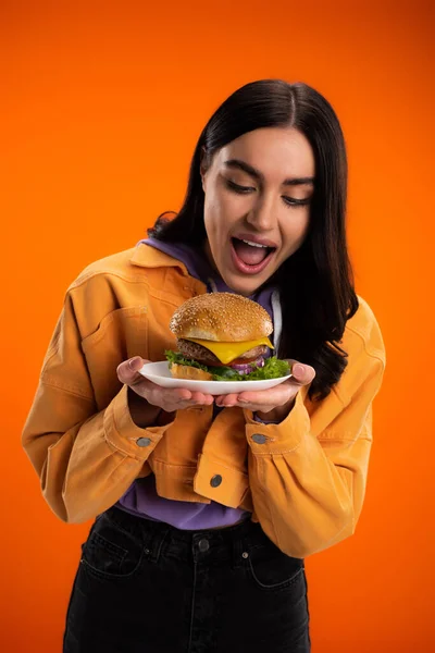 Hungry woman opening mouth near plate with delicious burger isolated on orange — Stock Photo