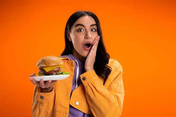 Вражена жінка в модній куртці тримає смачний бургер, торкаючись обличчя і дивлячись на камеру ізольовано на помаранчевому — стокове фото