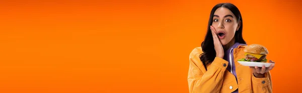 Изумленная женщина с вкусным бургером трогательное лицо и глядя на камеру изолированы на оранжевый, баннер — стоковое фото